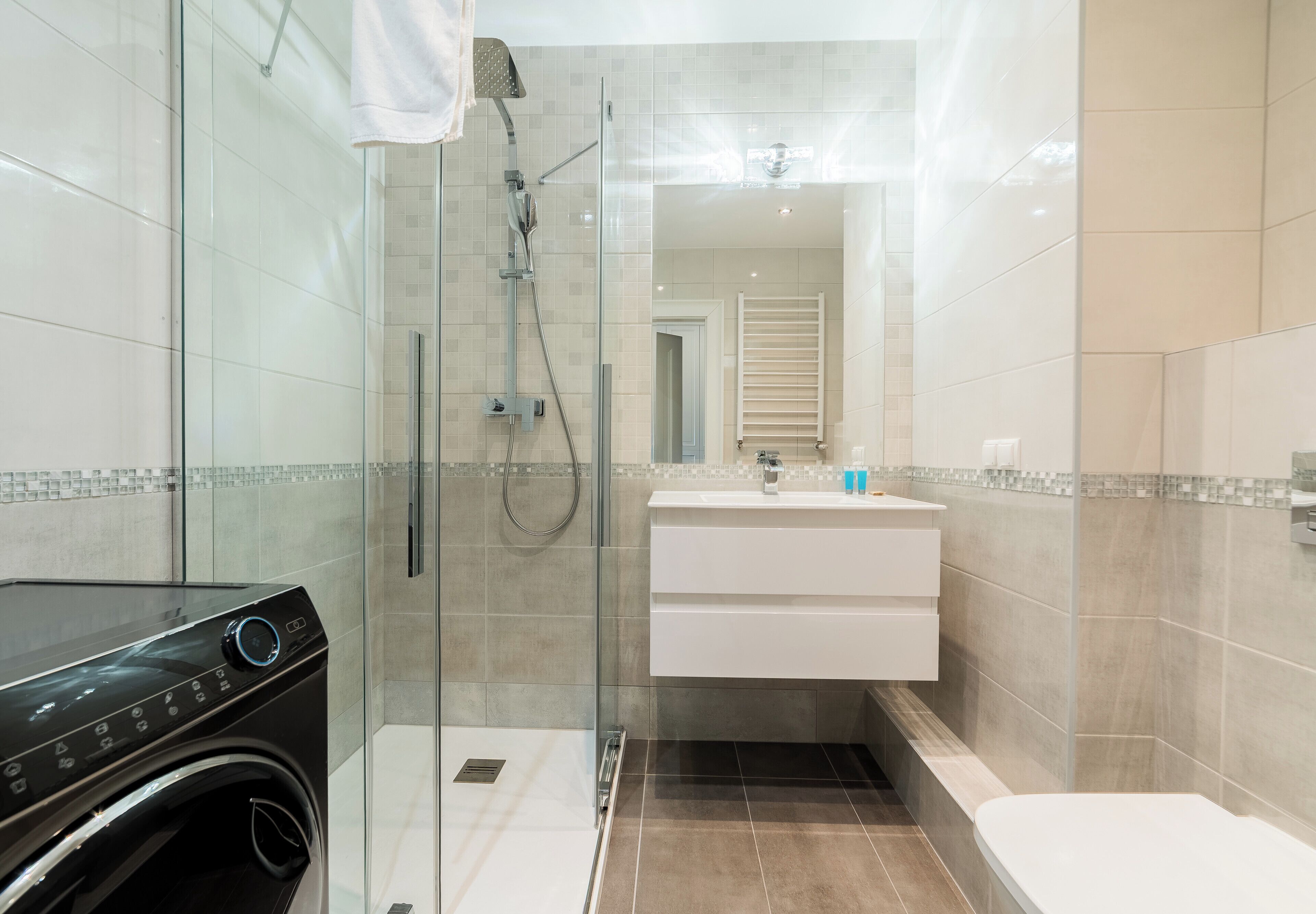 A modern bathroom with a glass-enclosed shower, a built-in washing machine, a white vanity with a mirror, and a toilet.