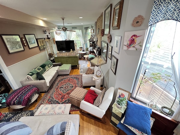 A cozy living room with a sofa, armchairs, a coffee table, and a TV. There are framed pictures on the walls, a ceiling fan, and a window with a view.