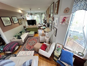 Un salon chaleureux avec un canapĂ©, des fauteuils, une table basse et un tĂ©lĂ©viseur. Les murs sont ornĂ©s de tableaux encadrĂ©s, il y a un ventilateur de plafond et une fenĂȘtre offrant une belle vue.
