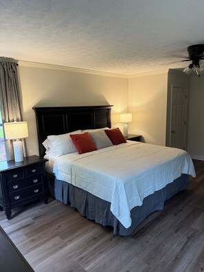 A neatly made bed with a white bedspread and multiple pillows, a dark wooden nightstand with a lamp, and a wooden floor. - Nana’s Cottage. Perfect 2 bedroom 1 bath home in a beautiful country setting (Attalla)