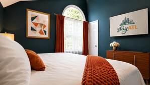 A hotel room with a large bed, a framed picture, a cityscape sign, and a window with curtains. - West Midtown | Hot Tub • Fire Pit • Parking (Atlanta)
