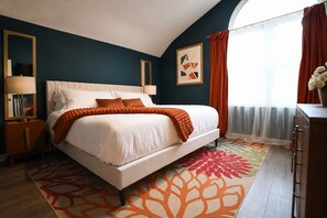 Une chambre à coucher comprenant un lit, des tables de chevet, une commode, une fenêtre avec des rideaux et un mur orné d’un tableau encadré.