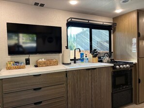A compact kitchen with a flat-screen TV, a window, a sink, and a stove. - Shave Lake Glamping Experience with King Bed near Peaceful Meadow. Walk to Town. (Shaver Lake)