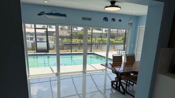 Et værelse med et stort vindue der kigger ud på en swimmingpool, en ventilator i loftet og et skilt med teksten 'BEACH HOUSE'.