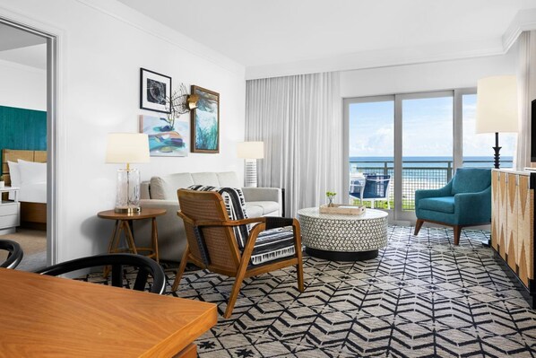 Ein Hotelzimmer mit Bett, rundem Couchtisch, einer Couch, einem Sessel, einem Fernseher und einem Balkon mit Meerblick.