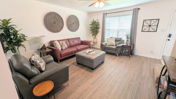 A living room with a sofa, armchair, ottoman, and a side table. There is a ceiling fan, a potted plant, and a window with blinds. - Sleep 6•Pet Friendly•Fully Stocked in East Bernard (East Bernard)