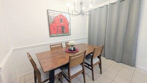 A dining area with a wooden table, four chairs, a chandelier, a painting of a red barn, and grey curtains.