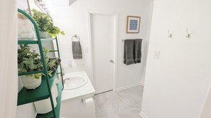 Une salle de bain avec un lavabo blanc, des étagères vertes ornées de plantes, une porte blanche et un tableau encadré au mur.
