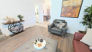 Un salon avec un canapé en cuir gris, une table basse ronde avec un plateau, une bibliothèque et, au fond, un coin repas.