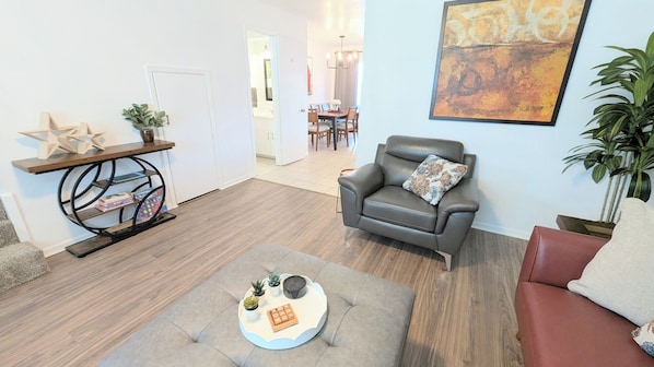 A living room with a grey leather sofa, a round coffee table with a tray, a bookshelf, and a dining area in the background.