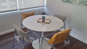 Ein runder, weißer Tisch mit einer Ananas als Mittelpunkt, umgeben von klaren Stühlen mit Leopardenmuster und gelben Kissen, in einem Raum mit einem großen Fenster und einer Pflanze auf dem Fensterbrett.