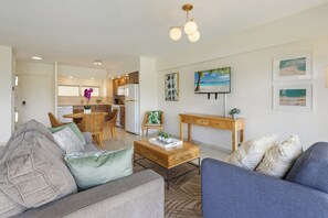 Ein modernes Wohnzimmer mit einer Couch, einem Couchtisch und einem blauen Sessel. Im Hintergrund ist eine Küche mit Essbereich zu sehen.