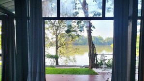Blick durch ein Fenster mit Vorhängen, einer Holzterrasse und einem See mit Bäumen.