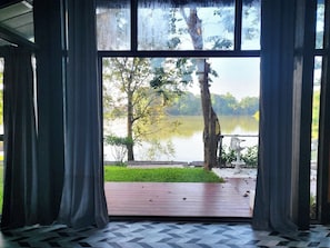 Vista a través de una ventana con cortinas, una terraza de madera y un lago rodeado de árboles.
