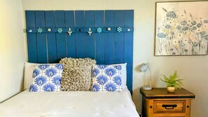 Ein Schlafzimmer mit Bett, einem Kopfende mit Meeresdekorationen, einem Nachttisch mit Schubladen und einem floralen Gemälde an der Wand.