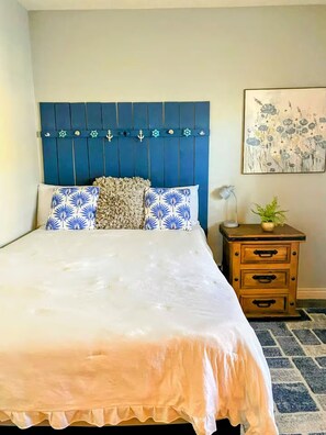 A bedroom with a bed, a headboard with nautical decorations, a nightstand with drawers, and a floral painting on the wall. - The little house (Bakersfield)