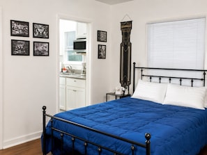 Un dormitorio con colcha azul, cabecera de metal negro, pared blanca con fotografías enmarcadas en blanco y negro, puerta blanca que da a una cocina y una ventana con persianas.
