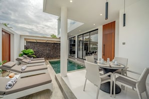 Un área de piscina moderna al aire libre con sillones de descanso, una mesa de comedor con sillas y un muro de piedra.