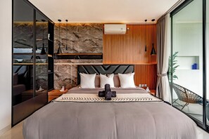 A modern bedroom with a large bed, a stone wall, wooden paneling, and a balcony with a chair. - Sky Beach Villa by Betterplace (Canggu)