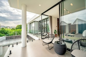 Un balcón moderno con barandas de vidrio, una zona de estar con sillas y una mesa, y vistas a áreas verdes y edificios.