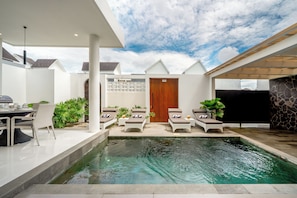 Un área de piscina exterior moderna con sillones reclinables, una mesa de comedor y un muro de piedra.