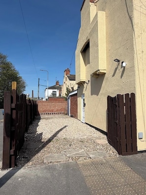 Un carrer estret amb un camí de grava que condueix a un edifici, amb una tanca de fusta d'un costat i una porta metàl·lica de l'altre.