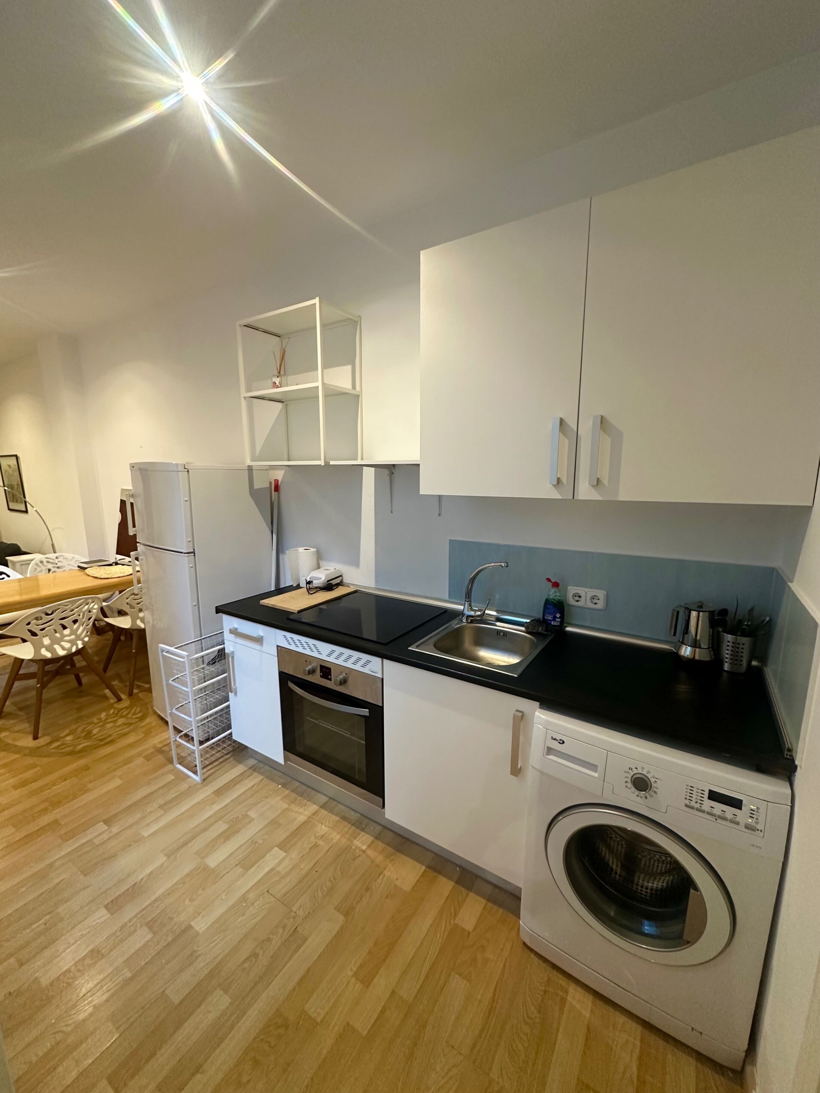 A compact kitchen with a dishwasher, oven, and washing machine, a dining area with a table and chairs, and a white cabinet with open shelves.