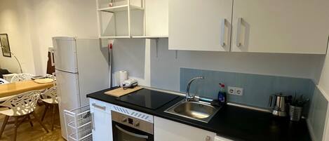 A compact kitchen with a dishwasher, oven, and washing machine, a dining area with a table and chairs, and a white cabinet with open shelves.