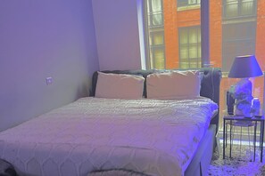 Ein Schlafzimmer mit Bett, Kissen, einem Nachttisch, einer Lampe und Blick auf ein Backsteingebäude durch ein Fenster.