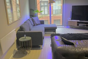 Ett modernt vardagsrum med en grå soffa, en svart läderbäddsoffa, ett litet runt sidobord, en platt-TV och ett stort fönster med utsikt över en byggnad med tegelstensfasad.
