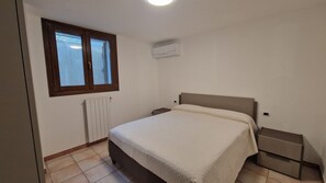 A bedroom with a bed, a nightstand, a window with blinds, a radiator, and an air conditioning unit. - Vrbo Property (Porto Frailis)