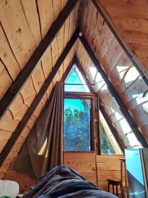 A wooden attic room with a sloped ceiling, a window with curtains, a bed, and a small table.