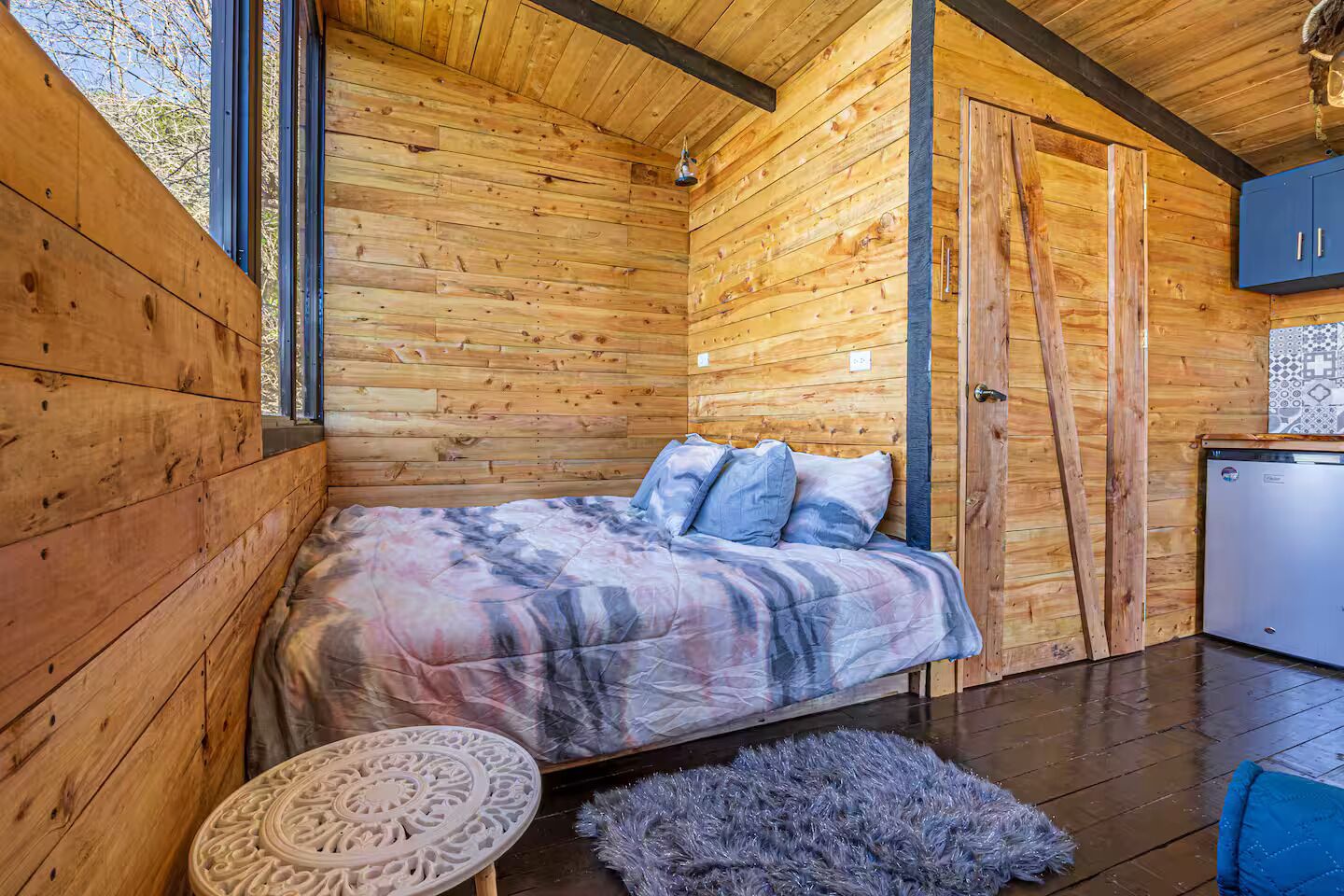 A wooden cabin interior with a bed, a small round table, a blue chair, and a kitchenette.