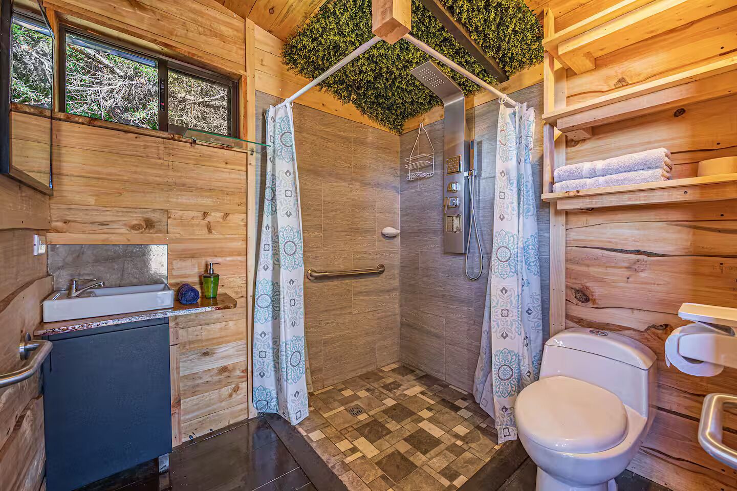 Une salle de bain rustique, aménagée dans une cabane en bois, avec une douche, des toilettes, un lavabo et une fenêtre donnant sur la forêt.