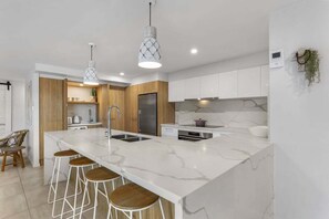 A modern kitchen with a marble island, bar stools, pendant lighting, and a built-in oven. - 3 Sundeck Gardens 72 Alexandra Parade (Maroochydore)