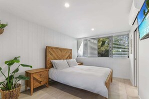 A bedroom with a bed, wooden headboard, white walls, a window with blinds, and a TV mounted on the wall. - 3 Sundeck Gardens 72 Alexandra Parade (Maroochydore)