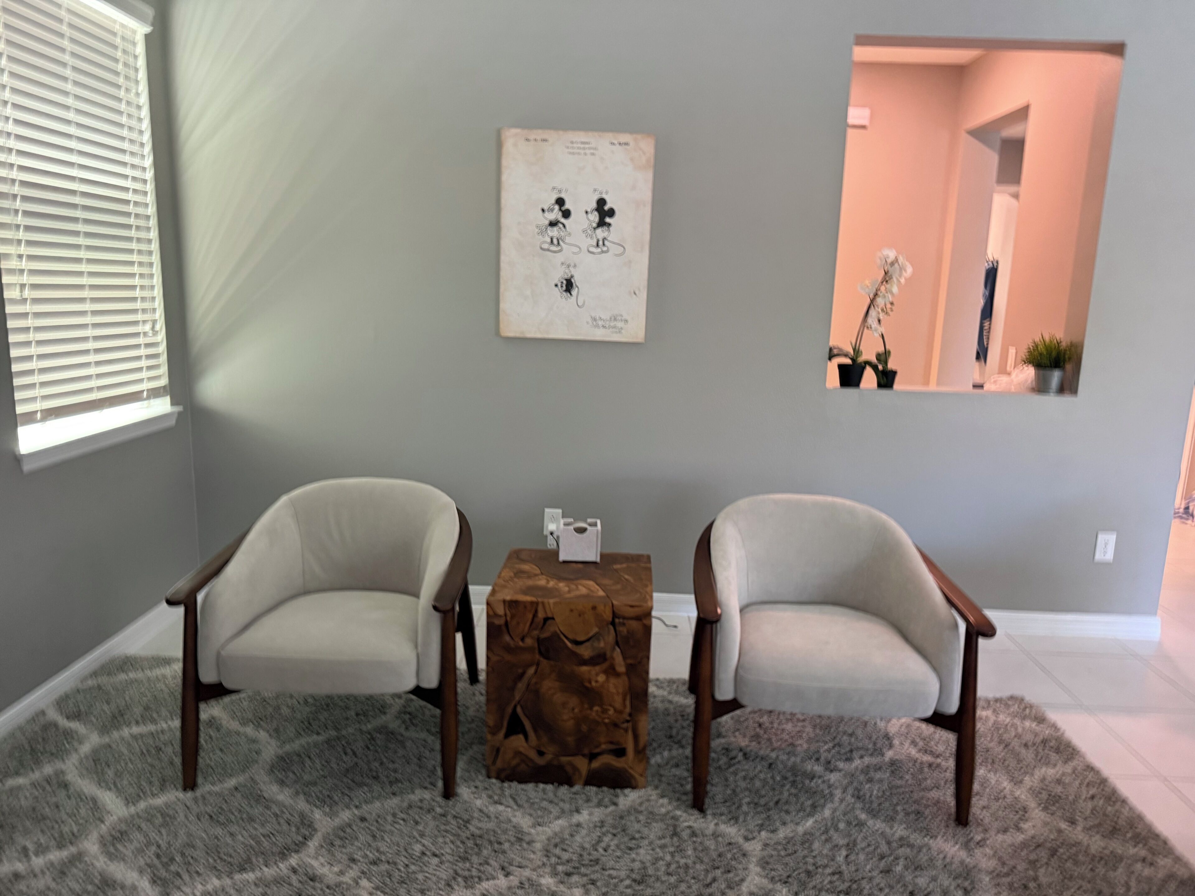A room with two armchairs, a small wooden table, a framed picture on the wall, and a window with blinds.