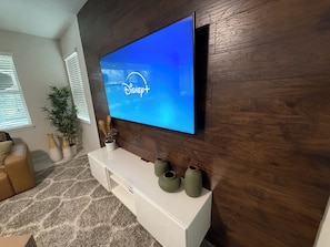 A living room with a flat-screen TV mounted on a wooden wall, a white entertainment unit with vases, and a potted plant. - Sleeps 13 - Luxurious - Heated Pool - Huge 3500sqft - Themed - Solara Resort! (Kissimmee)