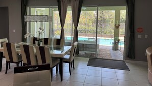 Un coin repas avec une table et des chaises en verre, un luminaire suspendu et une vue sur une piscine, visible à travers de grandes fenêtres.
