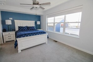Quarto com cama branca, paredes azuis, uma janela grande e um ventilador de teto.