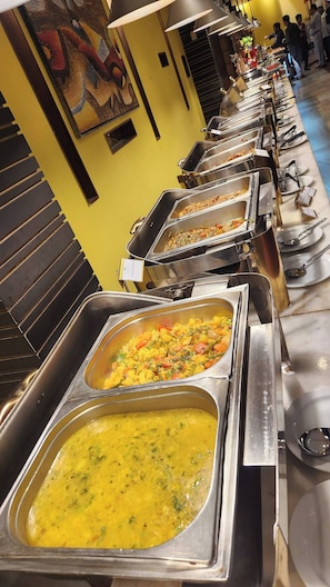 A buffet setup with multiple food stations, a yellow wall, and a painting. - Rose View Hotel (Sylhet)
