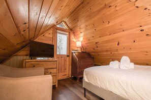 Un dormitorio acogedor en el ático con techo de madera, un televisor sobre un aparador, una lámpara y toallas ordenadas sobre la cama.
