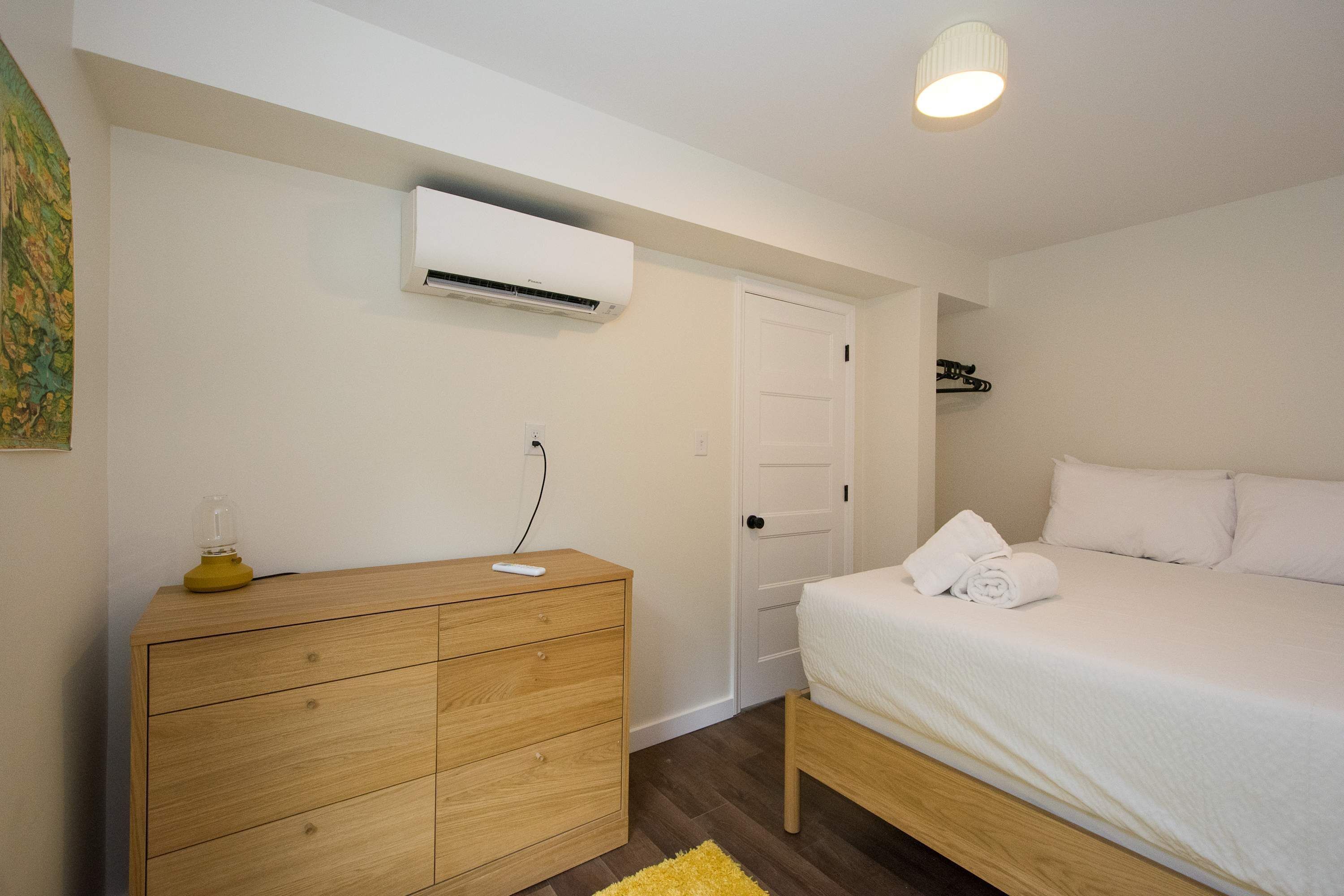 Une chambre à coucher comprenant un lit, une commode en bois, un appareil de climatisation et une porte.