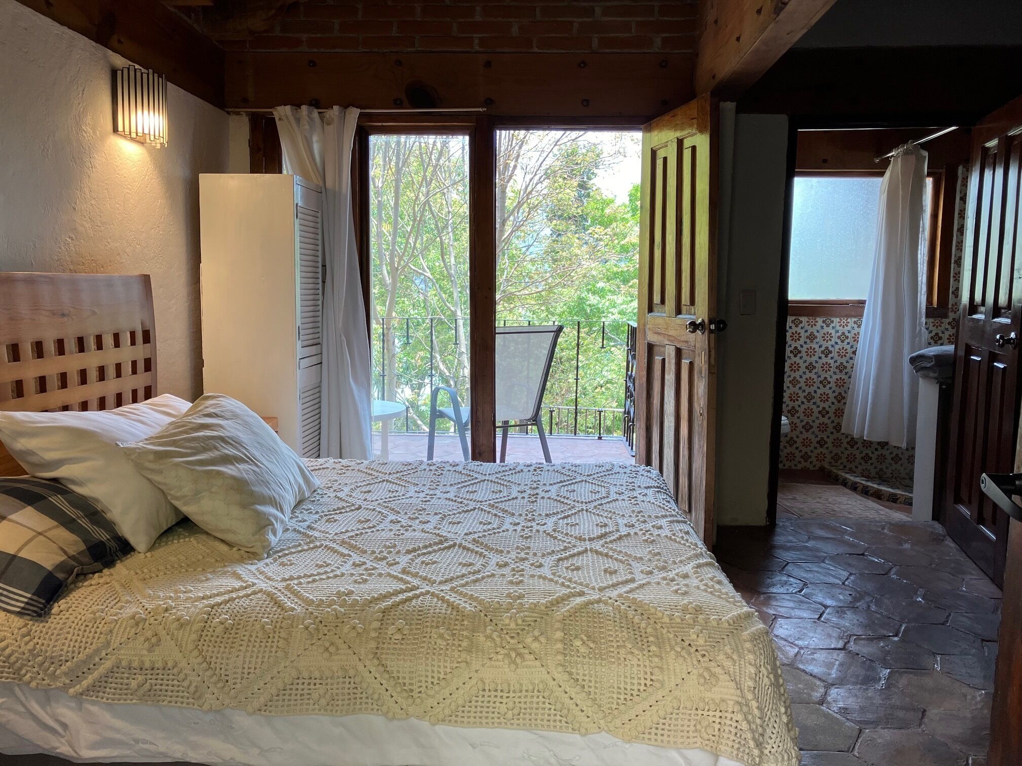 A bedroom with a large bed, a wooden headboard, a balcony with a chair, and a view of trees.