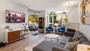 Ein modernes Wohnzimmer mit einer Couch, Sesseln, einem Couchtisch und einem Fernseher. Außerdem ein Bücherregal, ein Kamin und ein großes Fenster.