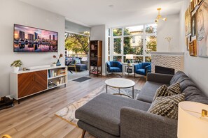 Ein modernes Wohnzimmer mit einer Couch, Sesseln, einem Couchtisch und einem Fernseher. Außerdem ein Bücherregal, ein Kamin und ein großes Fenster.