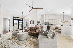 Ein geräumiges Wohnzimmer mit einem braunen Ledersofa, grauen Sesseln und einem Couchtisch. Eine Deckenleuchte und Pendelleuchten sind zu sehen.