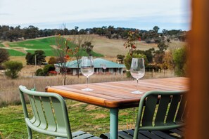 A table with two wine glasses and a chair, overlooking a rural landscape with fields and a building in the distance.