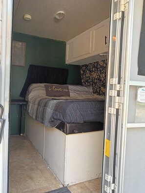 Interior compacto de una caravana con cama, una mesita y una puerta que da al exterior.