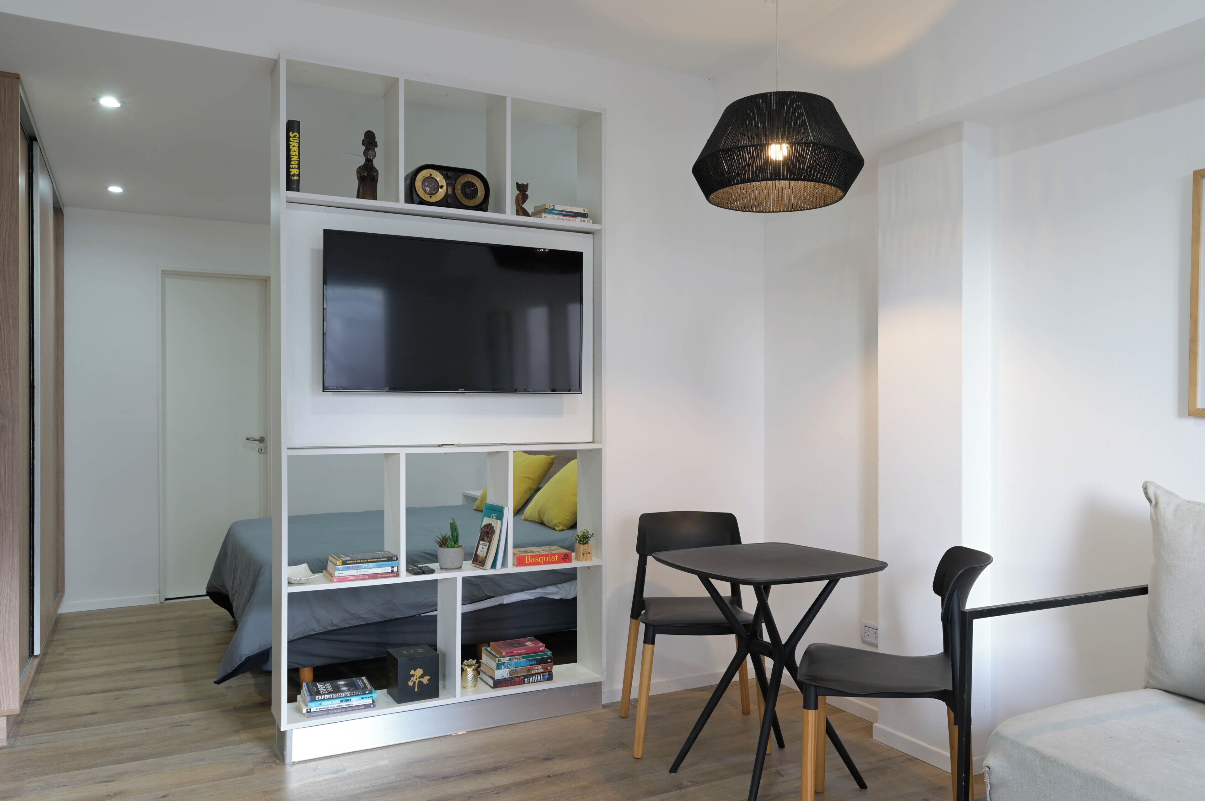 Una habitación moderna con cama, televisión montada en un mueble blanco, mesa de comedor con sillas y una lámpara colgante.